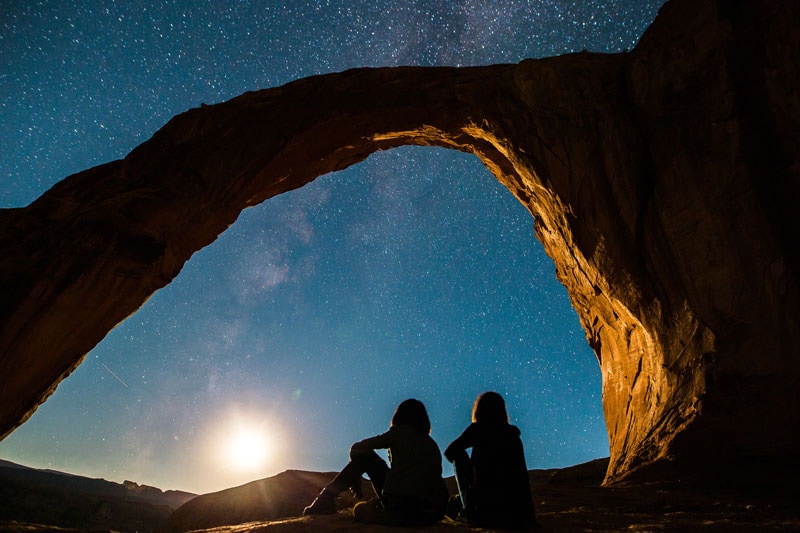 Two people sitting under a stone arch looking up at the night sky