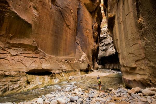 an orange stone slot canyon with a stream an two people in it